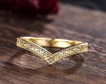 May include: A gold-colored, V-shaped ring with an intricate, engraved floral pattern. The ring is set against a dark wooden surface, highlighting its delicate design. The ring's band is approximately 2 mm wide.