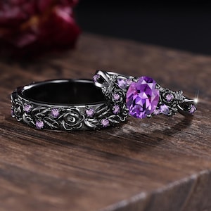 May include: Two black rings with floral and leaf designs. One ring features a large oval purple gemstone, while the other has smaller purple gemstones. The rings are set on a wooden surface.