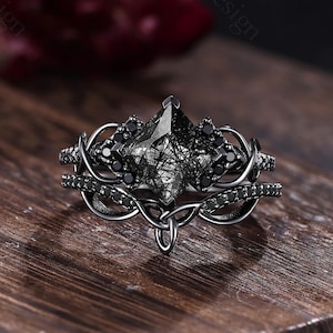 May include: A black metal ring set featuring a kite-shaped rutilated quartz center stone with black inclusions. The band and setting are adorned with small black stones and intricate, swirling metalwork that forms a decorative, vine-like pattern.