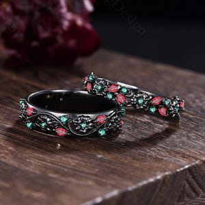 May include: Two black metal rings featuring an intricate floral pattern engraved along the bands. The rings are set with small, marquise-cut red stones and round, brilliant-cut green stones embedded within the dark metal floral design.