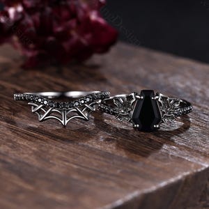 May include: A two-piece ring set featuring a black metal band with a coffin-shaped black gemstone flanked by leaf-like metalwork, and a matching curved band with a spiderweb design and small black stones set along the top edge of the band.