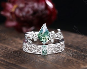May include: A silver engagement ring set with a green moss agate gemstone and a delicate leafy design. The ring is set on a wooden surface.