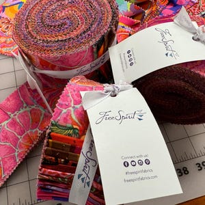 Op de afbeelding: Een close-up van stofrollen met roze en oranje patronen. De rollen zijn gelabeld met "Free Spirit" en hebben een wit lint eromheen geknoopt. De stof is geschikt voor quilten en knutselen.