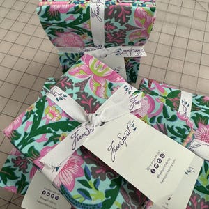 Puede incluir: Montones de paquetes de tela floral atados con cintas blancas y etiquetas. La tela presenta un patrón colorido con flores y hojas rosas, verdes y azules. Las etiquetas dicen "Free Spirit Fabrics" e incluyen iconos de redes sociales.