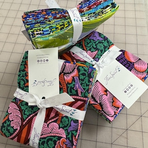 May include: Bundles of colorful fabric squares, tied with white ribbon and tags. The fabric features floral and abstract patterns in shades of green, pink, orange, and blue. The tags read "Free Spirit".
