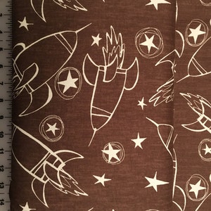 Peut inclure: Tissu brun avec un motif blanc répété de fusées et d'étoiles. Le tissu est étiqueté "Jeans and Things by David".