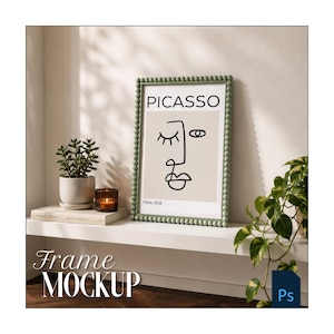 May include: A rectangular picture frame with a sage green beaded border containing a print of a Picasso line drawing of a face. The text PICASSO and Paris, 1928 are visible on the print. The frame is displayed on a white shelf next to a small potted succulent and a candle.