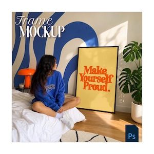 Peut inclure: Maquette d'affiche encadrée avec le texte "Make Yourself Proud" en orange sur fond jaune. Le cadre est noir. La personne porte un sweat-shirt bleu.