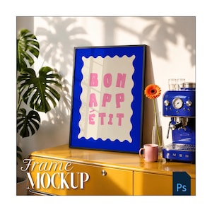 Pode incluir: Uma moldura retangular preta contendo uma impressão com uma borda creme ondulada sobre um fundo azul profundo. A impressão apresenta o texto rosa "BON APPÉTIT" em uma fonte sem serifa em negrito. A moldura está sobre um móvel amarelo.