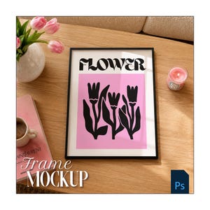 May include: A black rectangular frame containing a print with the word FLOWER in black letters above a pink square featuring three black tulip illustrations. The frame is positioned on a wooden surface next to a vase of pink tulips and a candle.
