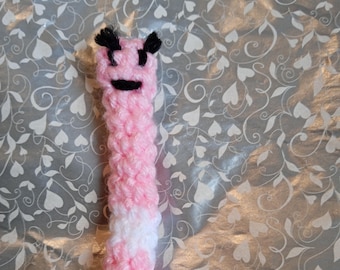 May include: A pink and white crocheted worm with a black smiling face and black eyes. The worm is on a white background with a heart pattern.