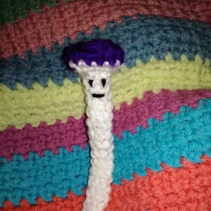 May include: A crocheted mushroom with a purple cap and a white stem. The mushroom has a smiling face.