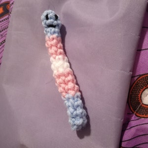 Puede incluir: Un gusano de ganchillo hecho a mano con los colores de la bandera del orgullo transgénero: azul claro, rosa y blanco. El gusano tiene una cara sonriente con ojos y boca negros. El gusano está sobre una superficie lavanda.
