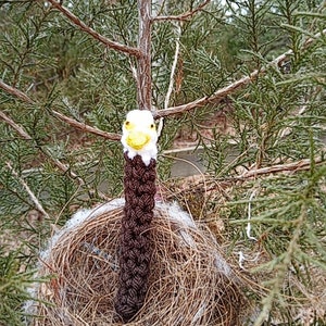 May include: A crocheted bald eagle figurine with a yellow beak and white head perched in a bird's nest made of twigs and straw.