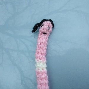May include: A pink crocheted doll with a white stripe and black hair. The doll has a simple face with two black eyes.