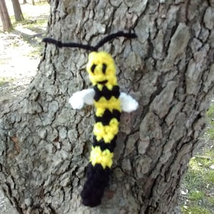 May include: A crocheted bee toy with a yellow and black striped body, white wings, and a black antenna. The bee has a smiling face with black eyes.
