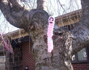 May include: A pink and white crocheted worm with a black face hanging from a tree branch. The tree is in front of a house with a red brick wall.