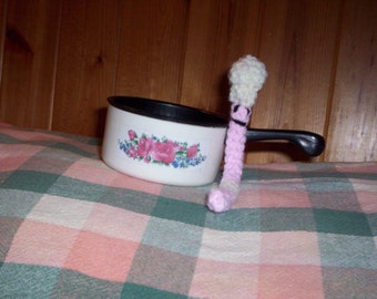 May include: A white ceramic saucepan with a black handle and floral design sits on a pink and green checkered tablecloth. A pink and white crocheted worm with a smiling face is next to the saucepan.