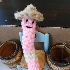 May include: A pink and white crocheted worm wearing a brown crocheted hat sits on a blue chair with a woven straw seat. The worm has a smiling face with black eyes. The worm is sitting in front of two brown ceramic pots.