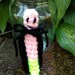 May include: A handmade crochet doll with a pink head, black eyes, and a smiling mouth. The doll has a black bow and a black body that fades into a green and yellow tail. The doll is placed in front of a glass jar.
