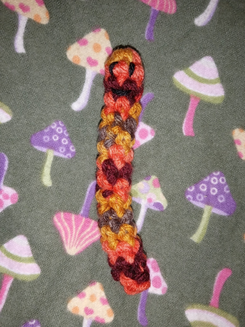 May include: A handmade, colorful crochet caterpillar toy. The caterpillar is made of orange, yellow, brown, and red yarn, with a smiling face. The background is a green fabric with a pattern of various mushroom designs.