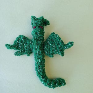 May include: A green crocheted dragon with silver glitter accents and red eyes. The dragon is curled up in a flying position.
