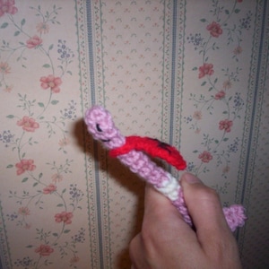 May include: A pink crocheted worm with a red cape, held in a hand against a floral wallpaper background.