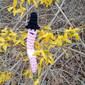May include: A pink and white crocheted worm with a black top hat. The worm is surrounded by yellow flowers.