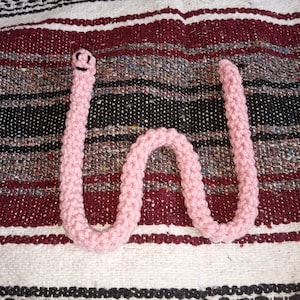 May include: A pink crocheted worm with a smiling face, lying on a striped blanket.