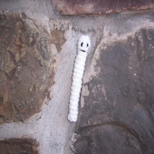 May include: A handmade white crochet worm with a smiling face. The worm is positioned against a textured stone wall, creating a whimsical contrast. The crochet worm is a unique decorative item.