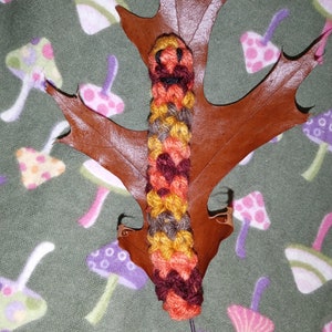 May include: A handmade, colorful crochet caterpillar bookmark. The caterpillar is made of orange, yellow, brown, and burgundy yarn. It rests on a brown leaf, with a green background featuring mushroom patterns.