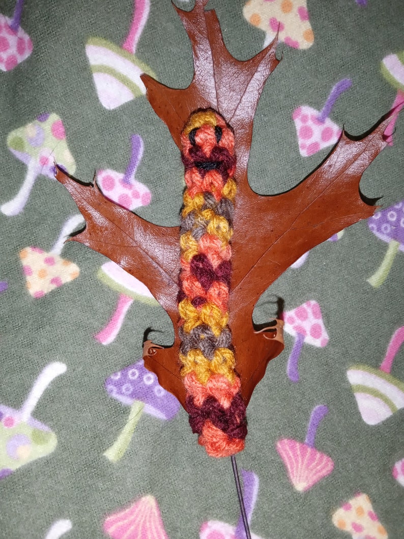 May include: A handmade, colorful crochet caterpillar bookmark. The caterpillar is made of orange, yellow, brown, and burgundy yarn. It rests on a brown leaf, with a green background featuring mushroom patterns.