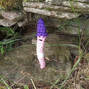 May include: A handmade crochet mushroom with a purple cap and a pink and white stem. The mushroom has a smiling face and is set against a backdrop of stone and grass. The whimsical design is a unique decorative item.
