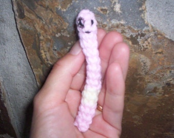 May include: A pink crocheted worm with a white band and black eyes. The worm is held in a person's hand.