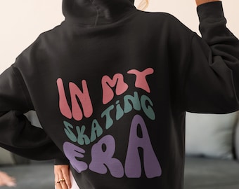 以下が含まれることがあります： 背面に「In My Skating Era」というカラフルなレトロな文字で書かれた黒いフード付きスウェットシャツです。