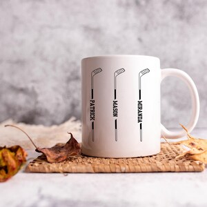May include: White ceramic mug featuring three black hockey stick graphics with the names "Patrick", "Mason", and "Miranda" printed vertically. The mug sits on a woven coaster with dried leaves in the foreground.