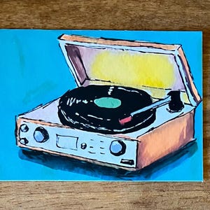May include: A hand-painted illustration of a portable record player with a tan casing and a black vinyl record on the platter. The turntable is depicted against a bright cyan background, featuring a yellow interior lid and a black tonearm.