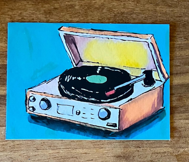 May include: A hand-painted illustration of a portable record player with a tan casing and a black vinyl record on the platter. The turntable is depicted against a bright cyan background, featuring a yellow interior lid and a black tonearm.