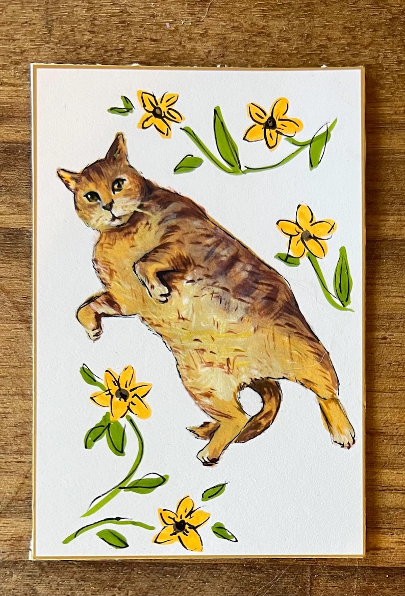 May include: A rectangular art print on white paper featuring a brown tabby cat lying on its side. The cat is surrounded by five hand-painted yellow flowers with green stems and leaves. The illustration uses visible brushstrokes and black ink outlines.