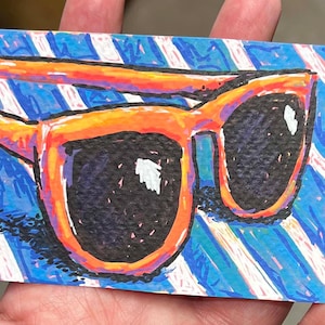 May include: A rectangular art print featuring a vibrant illustration of orange-rimmed sunglasses with dark lenses. The background consists of bold, diagonal blue and white stripes created with visible, textured crayon or marker strokes.