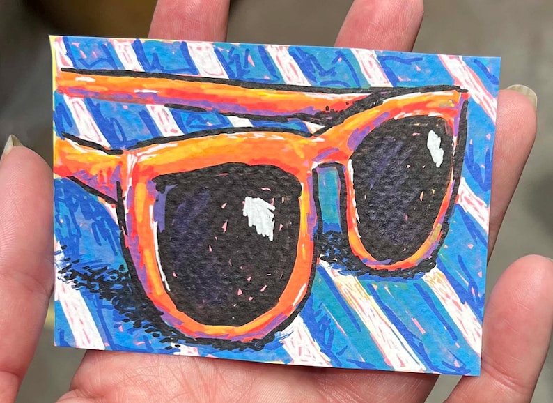 May include: A rectangular art print featuring a vibrant illustration of orange-rimmed sunglasses with dark lenses. The background consists of bold, diagonal blue and white stripes created with visible, textured crayon or marker strokes.