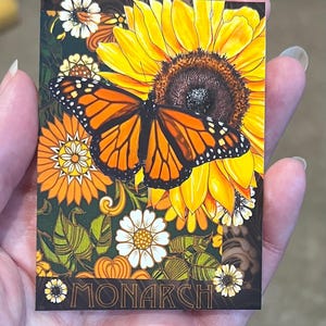 May include: A rectangular card featuring a vibrant orange and black monarch butterfly resting on a large yellow sunflower. The background includes smaller white and orange flowers, green leaves, and the word MONARCH printed in a bold font at the bottom.