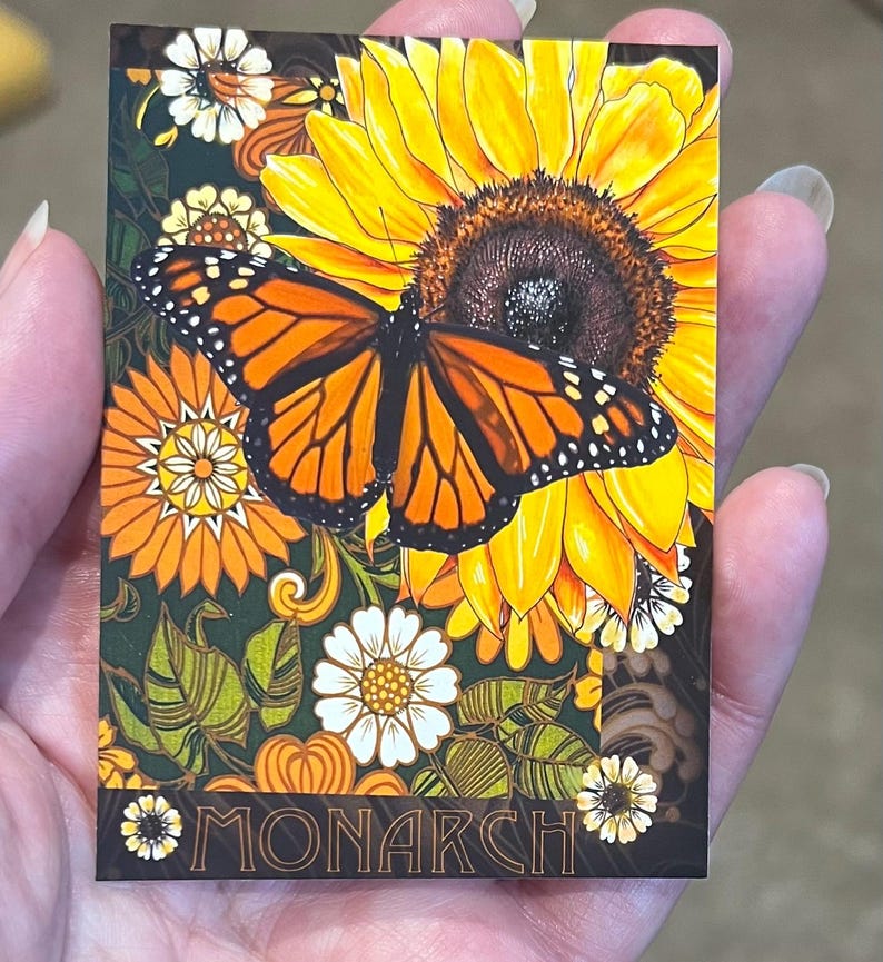 May include: A rectangular card featuring a vibrant orange and black monarch butterfly resting on a large yellow sunflower. The background includes smaller white and orange flowers, green leaves, and the word MONARCH printed in a bold font at the bottom.
