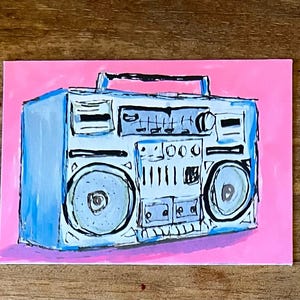 May include: A hand-painted illustration of a light blue boombox on a bright pink background. The boombox features a top handle, two large circular speakers, a central control panel with knobs and buttons, and a cassette deck area with black outlines.