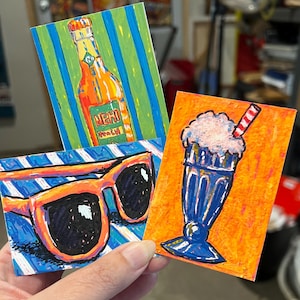 May include: Three rectangular art prints featuring vibrant illustrations. One shows an orange Nehi Peach soda bottle on a green and blue striped background. Another depicts orange-framed sunglasses on blue and white stripes. The third shows a blue milkshake.