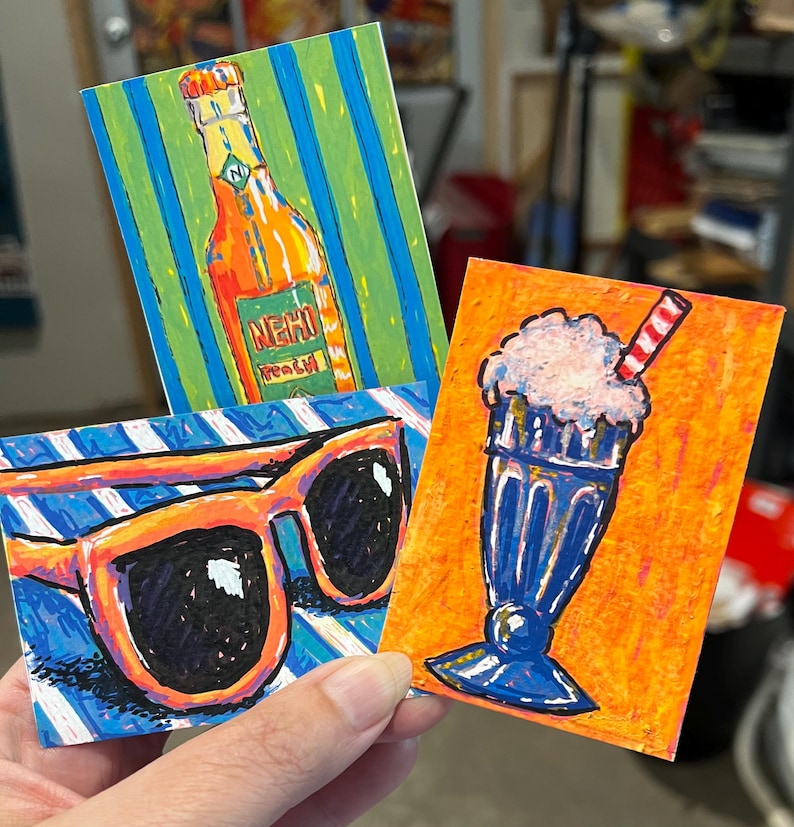 May include: Three rectangular art prints featuring vibrant illustrations. One shows an orange Nehi Peach soda bottle on a green and blue striped background. Another depicts orange-framed sunglasses on blue and white stripes. The third shows a blue milkshake.
