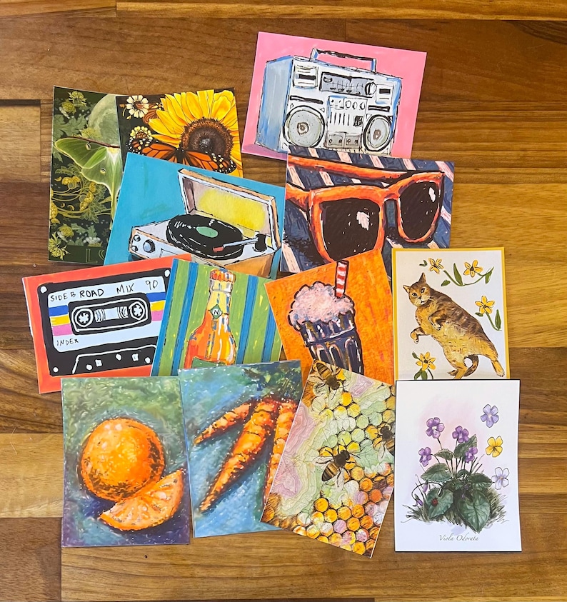May include: A collection of rectangular art prints featuring diverse illustrations including a boombox, sunglasses, a cassette tape labeled Side Road Mix 90, a record player, a cat, bees on a honeycomb, an orange, carrots, flowers, and a moth.