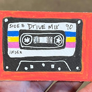 May include: A rectangular drawing of a black cassette tape with a bright orange border. The tape features blue, yellow, and pink horizontal stripes, with the handwritten text SIDE B DRIVE MIX 90 and the word INDEX visible on the white label area.