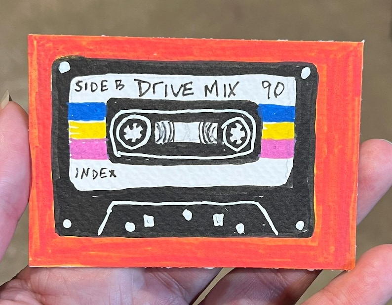 May include: A rectangular drawing of a black cassette tape with a bright orange border. The tape features blue, yellow, and pink horizontal stripes, with the handwritten text SIDE B DRIVE MIX 90 and the word INDEX visible on the white label area.
