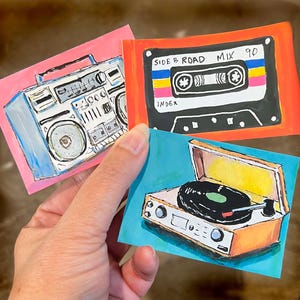 May include: Three rectangular stickers featuring hand-drawn illustrations of vintage audio equipment. One sticker shows a blue boombox on a pink background, another a black cassette tape labeled SIDE B ROAD MIX 90, and the third a record player.